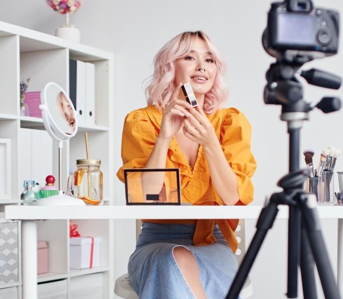 Full length, young beautiful blond Asian woman is recording make up tutorial video for beauty blog, wearing terracotta blouse. Video blogging, isolation, stay home concept