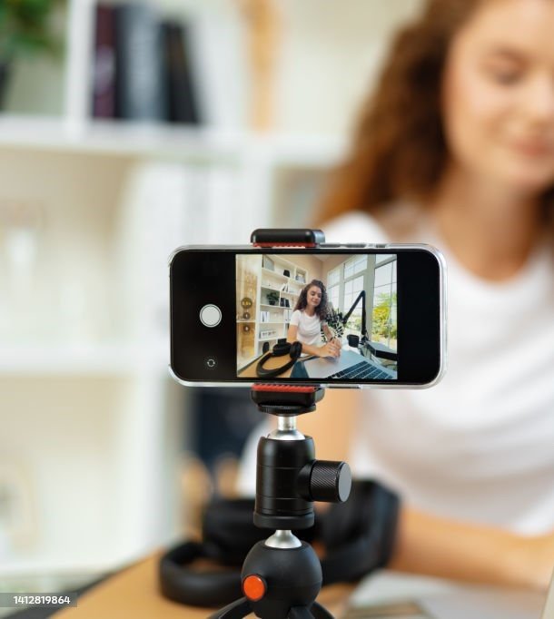 Young curly female blogger recording video at table at home on smartphone
